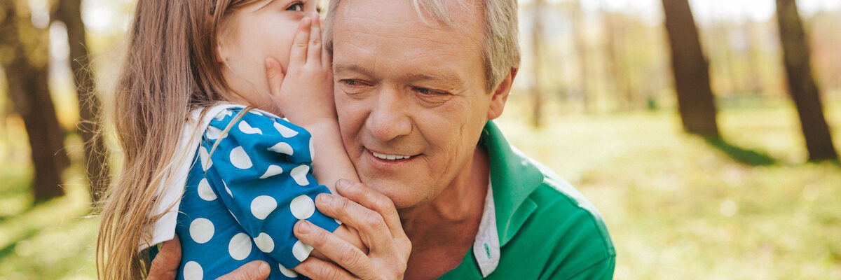 Adorable cute girl whispering to grandfather ear during the walk in park.